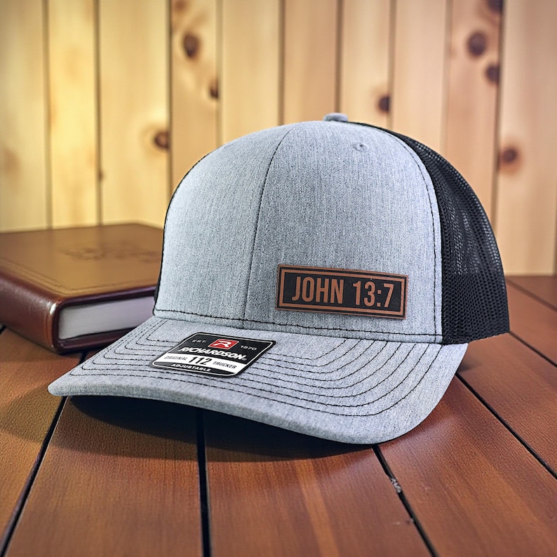 May include: A gray trucker hat with a black mesh back. The hat features a brown rectangular patch with the text "JOHN 13:7". The hat is sitting on a wooden surface.