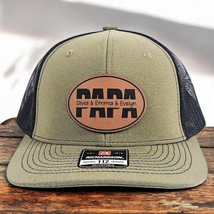 May include: A green and black trucker hat with a brown leather patch that says "Dada Olivia & Emma & Evelyn Papa". The hat is adjustable and has a Richardson logo.