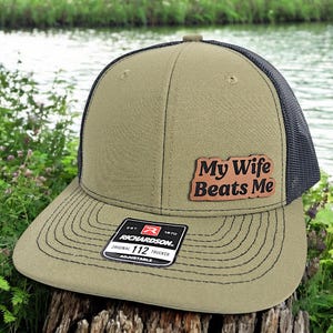 May include: A green and black trucker hat with a brown leather patch that says "My Wife Beats Me". The hat is adjustable and has a Richardson 112 logo.