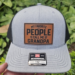 May include: A gray and black trucker hat with a brown leather patch that reads "My Favorite People Call Me Grandpa". The hat has a black mesh back and a Richardson logo with the text "Est. 1970 Richardson. Original 112 Trucker Adjustable".
