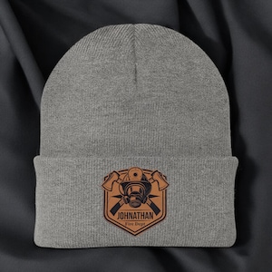 May include: A gray beanie with a brown leather patch featuring a firefighter's helmet, axes, and the text "JONATHAN Fire Dept." The beanie is made of a soft, knitted material, perfect for cold weather.