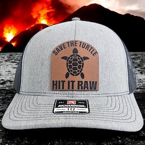 May include: A gray and black trucker hat with a brown leather patch that says "Save the Turtle" and "Hit it Raw". The patch features a black silhouette of a turtle. The hat is adjustable and has a Richardson logo.
