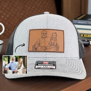 May include: A gray and black trucker hat with a leather patch featuring a sketch of a person and a child. The hat has a curved brim and a mesh back. The patch reads "RICHARDSON 112 TRUCKER ADJUSTABLE".