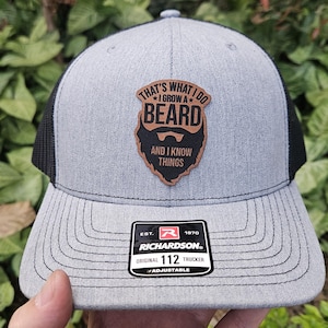 May include: A gray and black trucker hat with a brown leather patch that says "That's what I do *I grow a* BEARD and I know things". The hat is adjustable and has the Richardson logo on the side.