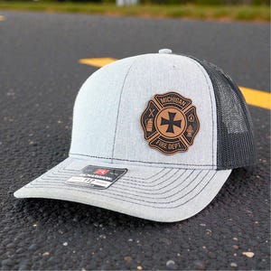 May include: A light gray and black trucker hat with a leather patch featuring the words "Michigan Fire Dept" and a Maltese cross emblem. The hat has a curved brim and a mesh back.