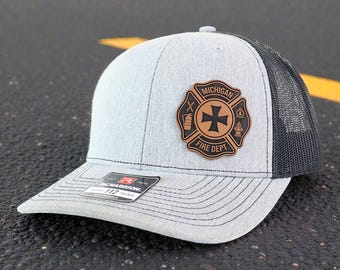 May include: A light gray and black trucker hat with a leather patch featuring the words "Michigan Fire Dept" and a Maltese cross emblem. The hat has a curved brim and a mesh back.