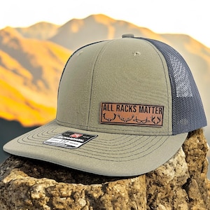 May include: A green and black trucker hat with a brown leather patch that reads "ALL RACKS MATTER" with a silhouette of antlers.