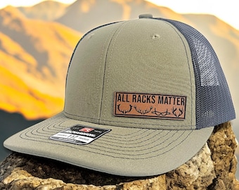 Puede incluir: Una gorra de camionero verde y negra con un parche de cuero marrón que dice "ALL RACKS MATTER" con una silueta de cuernos.