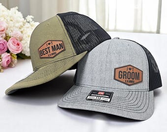 May include: Two trucker hats, one olive green and one gray, with black mesh backs. The olive hat has "BEST MAN" on a brown patch, and the gray hat says "GROOM". Both have a Richardson 112 Trucker tag.
