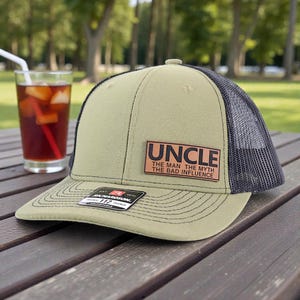 May include: Olive green trucker hat with a black mesh back. The hat features a brown leather patch with the word "UNCLE" and the phrase "THE MAN THE MYTH THE BAD INFLUENCE" in black text. A glass of iced tea sits next to the hat.