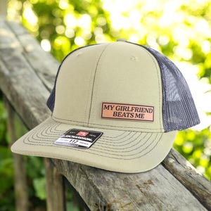 May include: Tan and navy blue trucker hat with the text "MY GIRLFRIEND BEATS ME" on a rectangular patch. The hat has a curved brim and a mesh back. The Richardson brand label is visible.