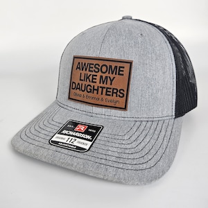 May include: Gray and black trucker hat with a brown leather patch that says "Awesome Like My Daughters Olivia & Emma & Evelyn". The hat has a black mesh back and a Richardson 112 label.