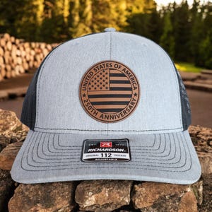 May include: A gray trucker hat with a black mesh back and a curved brim. The front features a circular patch with the American flag and the text "UNITED STATES OF AMERICA 250TH ANNIVERSARY". The hat includes a Richardson patch.