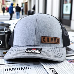 May include: Gray trucker hat with a black mesh back. The hat features a brown leather patch with Roman numerals and a Richardson patch. The hat is sitting on a newspaper.