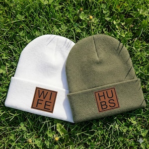 May include: Two beanies on green grass. One is white with a brown square patch that says "WIFE". The other is olive green with a brown square patch that says "HUBS".