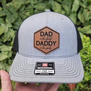 May include: A gray and black trucker hat with a brown leather patch that says "DAD in the streets DADDY in the sheets". The hat has a black mesh back and an adjustable strap with the Richardson logo and "112 TRUCKER" text.
