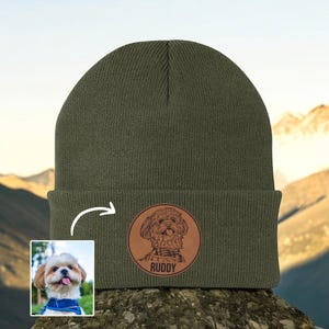 May include: Olive green beanie with a circular leather patch featuring a dog illustration and the name "Ruddy". A small photo of the dog is in the lower left corner. The beanie is displayed outdoors.