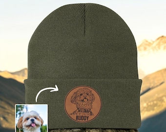 May include: Olive green beanie with a circular leather patch featuring a dog illustration and the name "Ruddy". A small photo of the dog is in the lower left corner. The beanie is displayed outdoors.