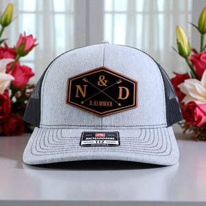 May include: A light gray trucker hat with a black mesh back and a curved brim. The front features a brown leather patch with the letters "N & D" and the Roman numeral date "X.XI.MMXX". The hat has an adjustable strap.