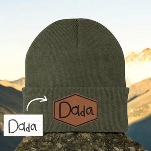 May include: Olive green beanie with a brown leather patch that reads "Dada" in a handwritten style. The beanie is made of a ribbed knit material. The background shows a mountain landscape.