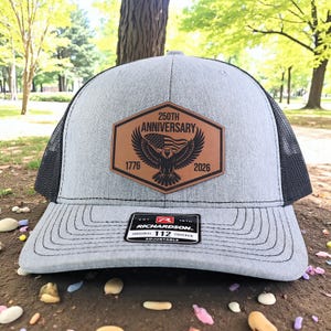 May include: A gray trucker hat with a black mesh back and a curved brim. The front features a brown leather patch with an eagle, American flag, and the text "250TH ANNIVERSARY 1776 2026". The hat has a Richardson 112 Trucker label.