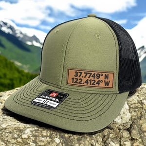 May include: Olive green trucker hat with a black mesh back. A brown leather patch displays the coordinates 37.7749° N 122.4124° W. The hat features a curved brim and an adjustable strap. The Richardson brand label is visible.