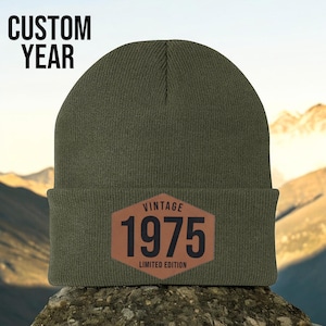 May include: An olive green beanie with a brown leather patch. The patch features the text "VINTAGE 1975 LIMITED EDITION". The words "CUSTOM YEAR" are at the top. The beanie is sitting on a rock with a mountain background.