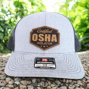 May include: A gray trucker hat with a black mesh back and a curved brim. The hat features a brown leather patch with the words "Certified OSHA Violator" and three stars. A black and white tag reads "Richardson Original 112 Trucker Adjustable".