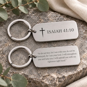 May include: Two rectangular silver-toned metal keychains with rounded corners and split rings. The top keychain features a black cross icon and the text ISAIAH 41:10. The bottom keychain is engraved with the full Bible verse from Isaiah 41:10.
