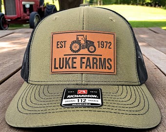 May include: Olive green and black trucker hat with a leather patch featuring a tractor graphic and the text "LUKE FARMS EST 1972". The hat has a curved brim and an adjustable fit.