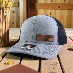 May include: A gray and black trucker hat with the text "FAITH CAN MOVE MOUNTAINS MATTHEW 17:20" on a brown patch. The hat has a curved brim and an adjustable strap.