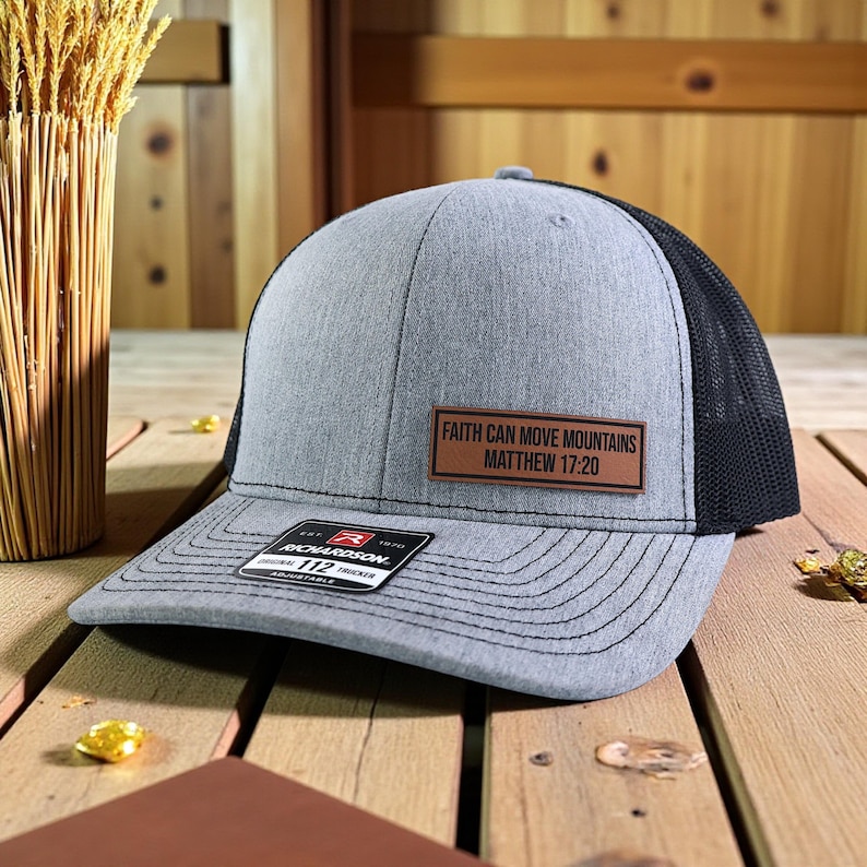 Puede incluir: Gorra de camionero gris y negra con el texto "FAITH CAN MOVE MOUNTAINS MATTHEW 17:20" en un parche marr&oacute;n. La gorra tiene una visera curva y una correa ajustable.