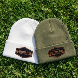 May include: Two beanies, one white and one olive green, rest on green grass. Each beanie has a brown patch with the word "PROBLEM" or "Somebody's PROBLEM" in white lettering. The beanies are folded at the brim.