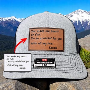 May include: Gray trucker hat with a black mesh back and a brown leather patch. The patch features the text: "You make my heart so full. I'm so grateful for you. With all my love, Sarah."