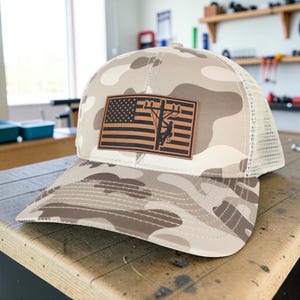 May include: A camouflage baseball cap with a brown and tan pattern. The cap features a tan mesh back and a brown leather patch with an American flag and a utility worker silhouette.