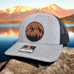 May include: A gray and black trucker hat with a brown leather patch featuring a silhouette of two mountains and trees. The hat is adjustable and has the text "Richardson 112" on the back.