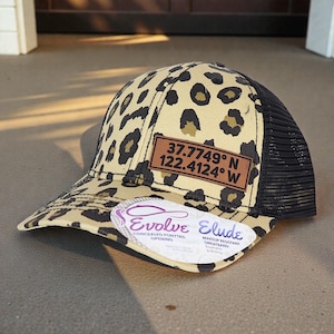 May include: A tan and black leopard print trucker hat with a black mesh back. The hat features a brown leather patch with the coordinates 37.7749° N 122.4124° W. A white tag with the text "Evolve Elude" is attached.