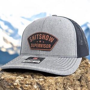 May include: A gray and black trucker hat with a brown leather patch that says "SHITSHOW SUPERVISOR" with three stars.