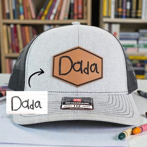May include: A gray and black trucker hat with a leather patch that says "Dada". The hat has a mesh back and a curved brim. The patch is brown with black lettering. The hat is adjustable.