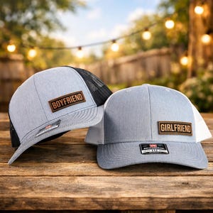 May include: Two gray trucker hats with the words "BOYFRIEND" and "GIRLFRIEND" on brown patches. One hat has a black mesh back, the other a white mesh back. Both have a Richardson 112 tag.