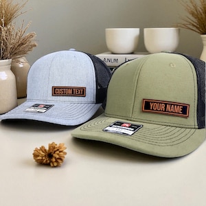 May include: Two trucker hats with mesh backs. One is light gray with "CUSTOM TEXT" on a rectangular patch, the other is olive green with "YOUR NAME" on a patch. Both have a curved bill and a black mesh back.