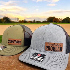 May include: Two trucker hats with leather patches. One is olive green with "SOMEBODY" in black. The other is gray with "Somebody's PROBLEM" in black. Both have black mesh backs and adjustable straps.