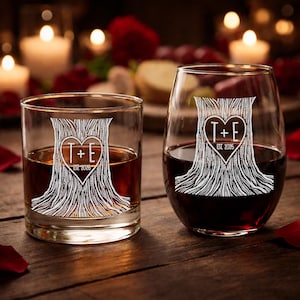 May include: Two clear glasses, one a rocks glass and the other a stemless wine glass, each etched with a tree design. The tree has a heart-shaped center with the initials "T+E" and the year "EST 2026". Both glasses are filled with liquid.