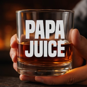 May include: A clear rocks glass filled with amber liquid, with the white text "PAPA JUICE" printed on the front. The glass is held by a person's hand, with a blurred background.