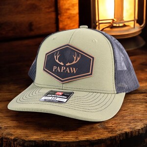 May include: A green and gray trucker hat with a black patch featuring the word "PAPAW" and deer antlers. The hat has a curved brim and a mesh back. The patch is a hexagon shape with a brown border.
