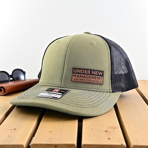 May include: Olive green and black trucker hat with the text "UNDER NEW MANAGEMENT - ASK WIFE FOR DETAILS" on a brown patch. The hat has a curved brim and a mesh back. Sunglasses and a brown wallet are in the background.