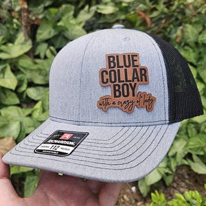 May include: A gray and black trucker hat with a brown wooden patch that says "BLUE COLLAR BOY with a crazy lady". The hat is adjustable and has a Richardson 112 trucker logo.