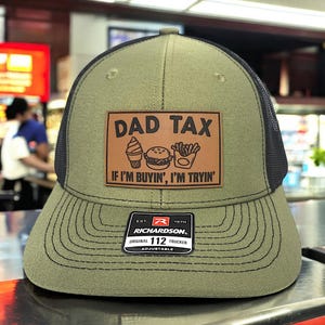 May include: Olive green trucker hat with a black mesh back and a brown leather patch. The patch reads "DAD TAX" above illustrations of an ice cream cone, burger, and fries, with the text "IF I'M BUYIN', I'M TRYIN'". The hat has a curved brim and an adjustable strap.