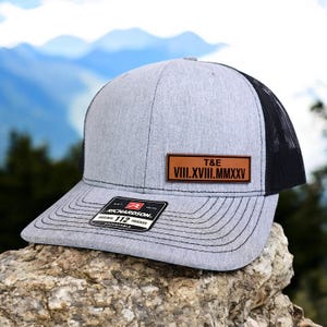 May include: Gray trucker hat with a black mesh back and a curved brim. A brown leather patch displays the initials "T&E" and Roman numerals. The hat has an adjustable strap and a "Richardson" tag.
