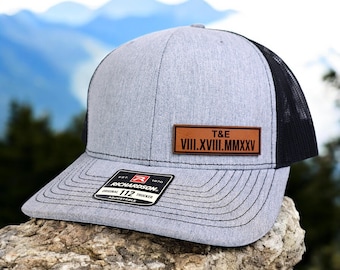 Peut inclure: Casquette de camionneur grise avec un dos en filet noir et une visière incurvée. Un patch en cuir marron affiche les initiales "T&E" et des chiffres romains. La casquette a une sangle réglable et une étiquette "Richardson".